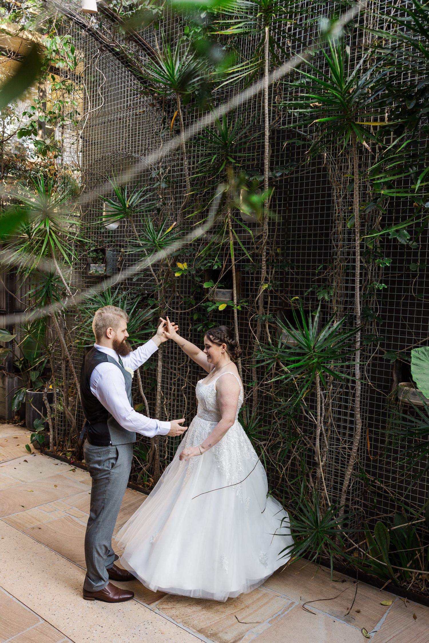 A bride and groom share a romantic dance outdoors surrounded by lush green tropical plants and a wire trellis wall.