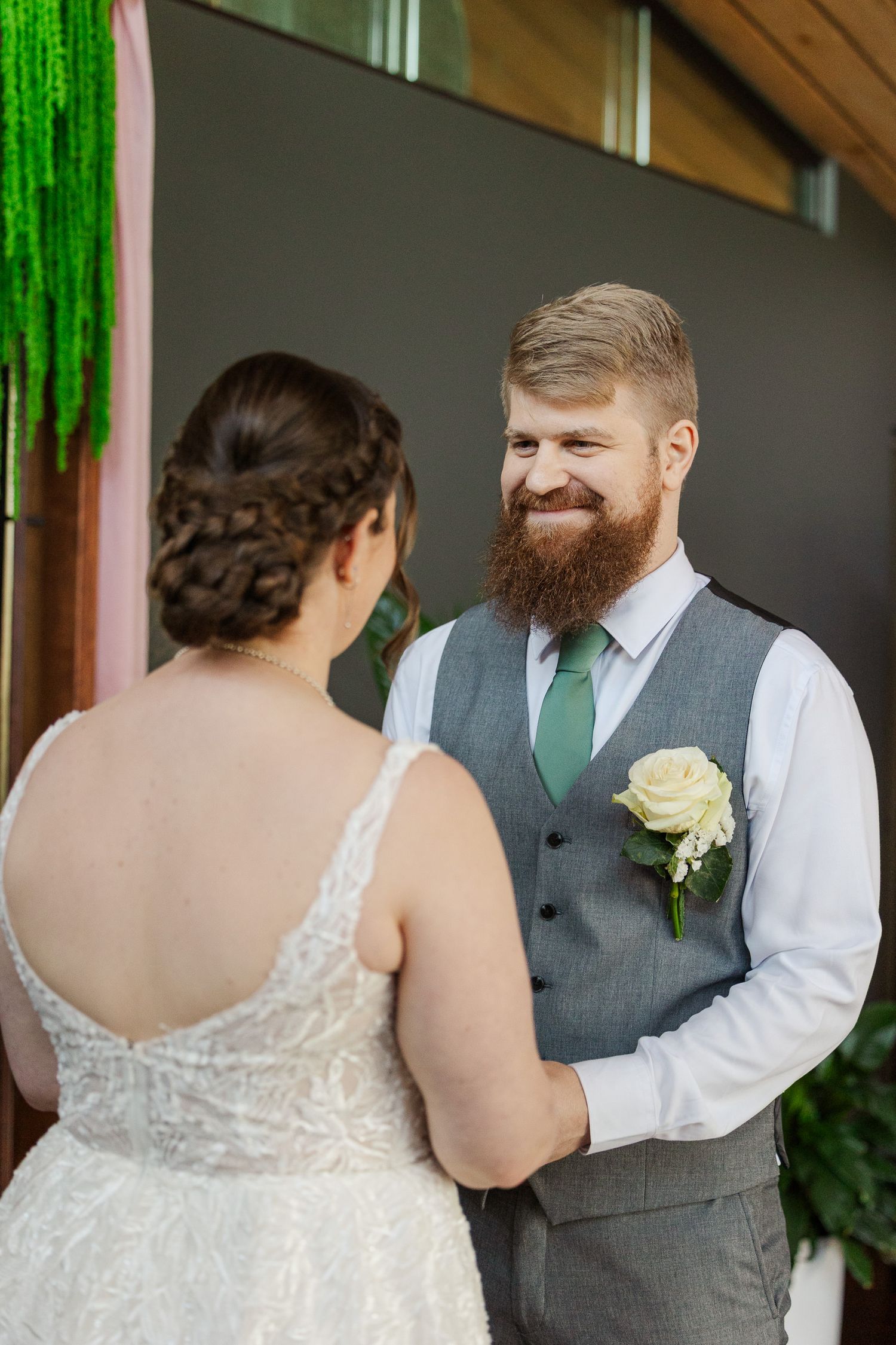 Bearded groom in gray vest and green tie smiles at bride during outdoor wedding ceremony with floral boutonniere.