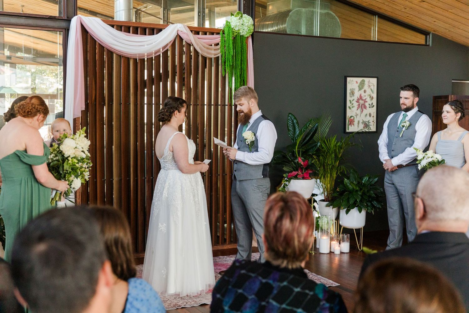 Bride and groom exchange vows at indoor wedding ceremony with greenery decor, bridesmaids, and groomsmen in soft sage tones.