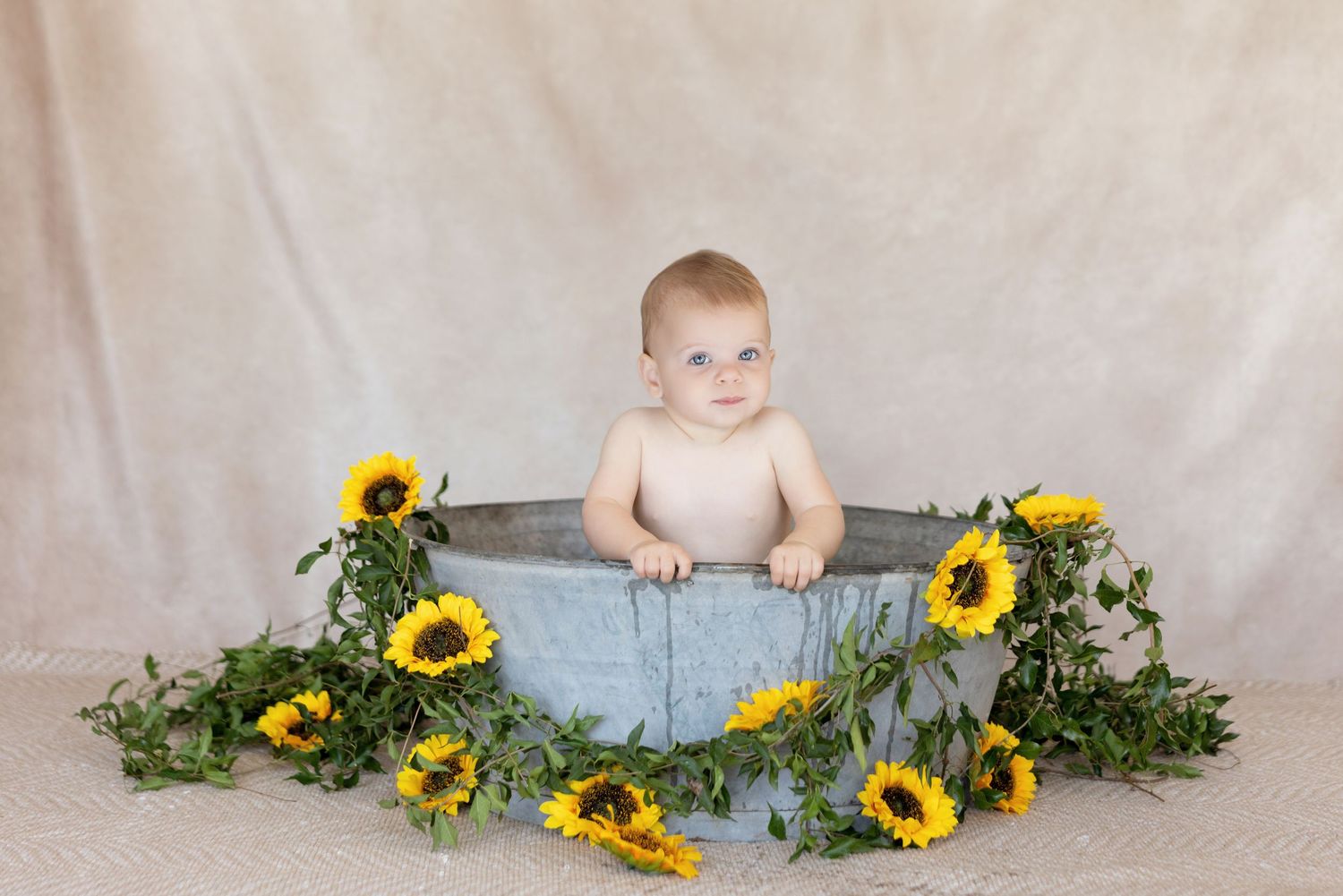 A baby sits in a blue metal tub surrounded by bright yellow sunflowers and greenery against a neutral backdrop.