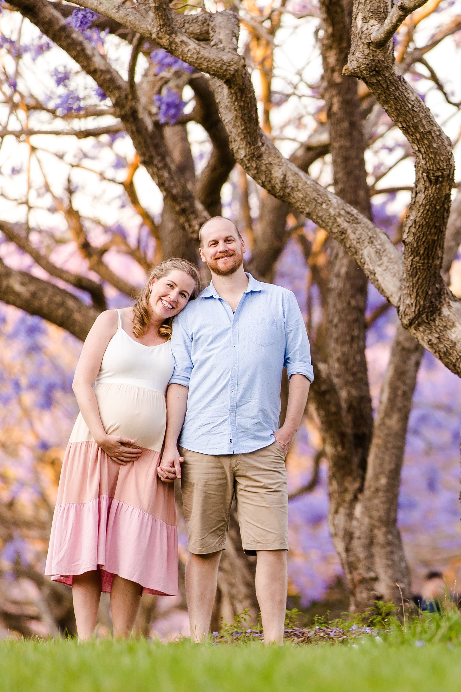 Group in light pink dresses and casual attire pose together under blooming jacaranda trees in a park setting.