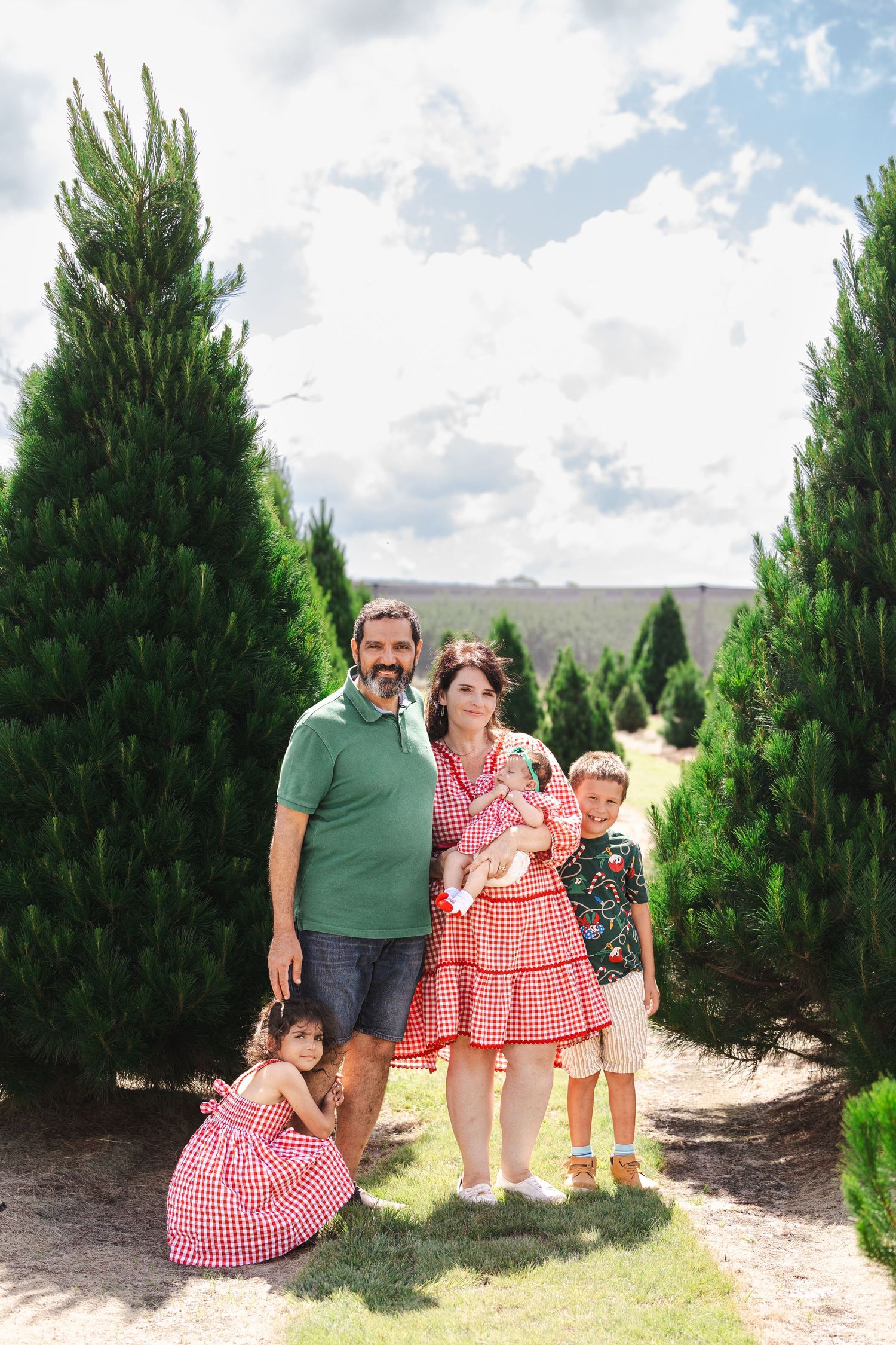 A family poses together between evergreen trees at a Christmas tree farm on a sunny day.