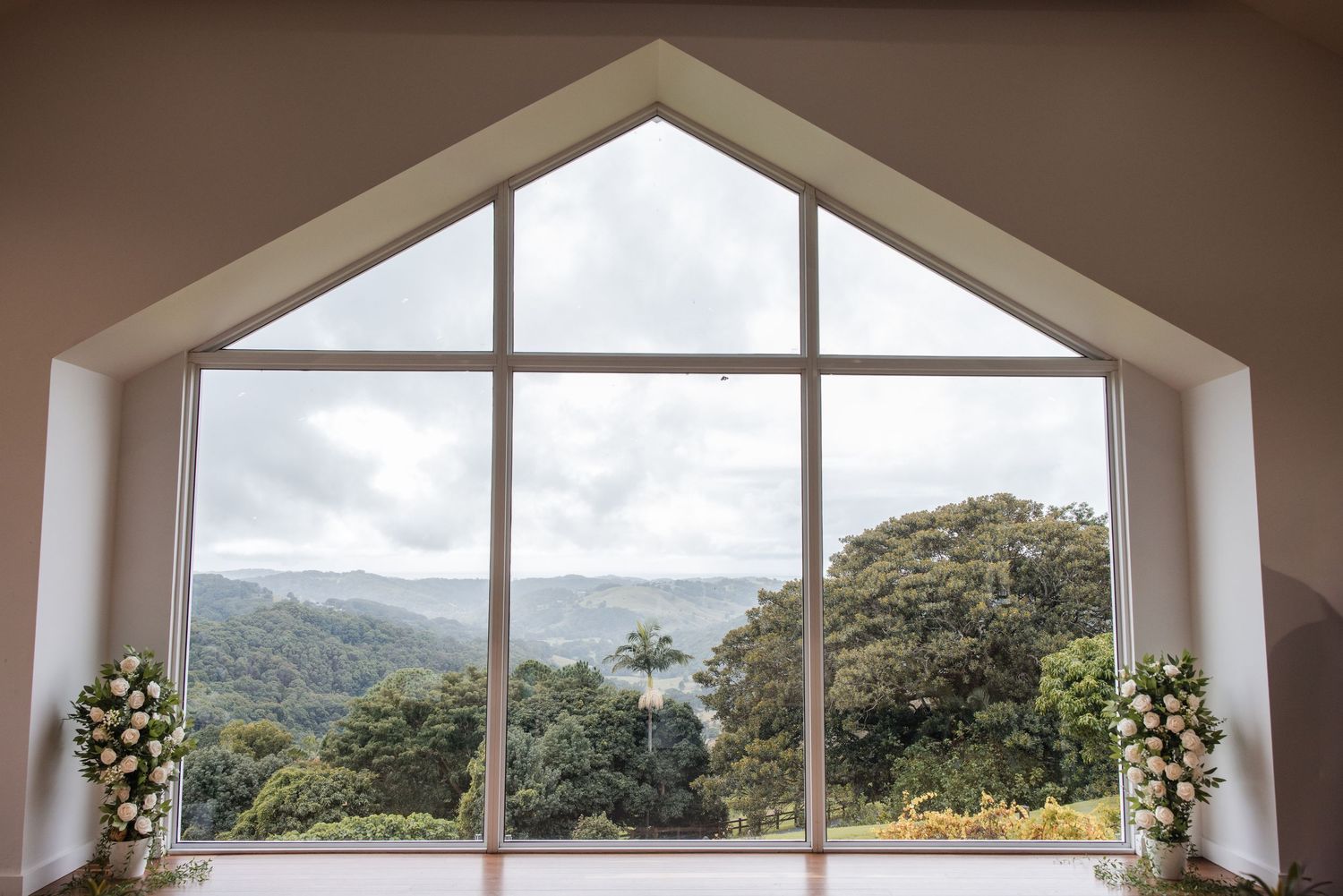 A-frame window with white trim overlooking lush green tropical trees and cloudy sky, flanked by white floral arrangements.