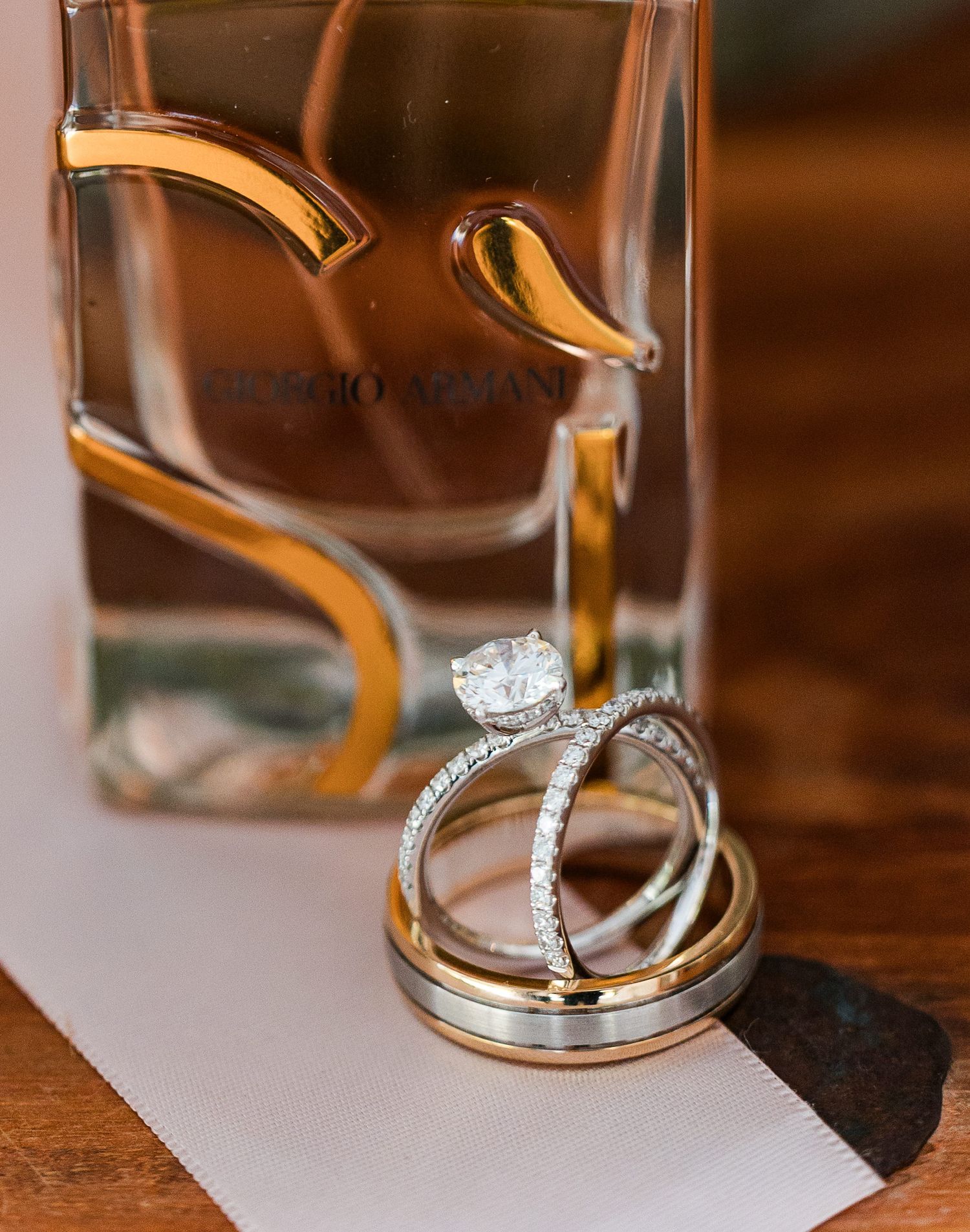 Wedding rings and diamond engagement ring stacked beside a Giorgio Armani perfume bottle on a wooden surface.