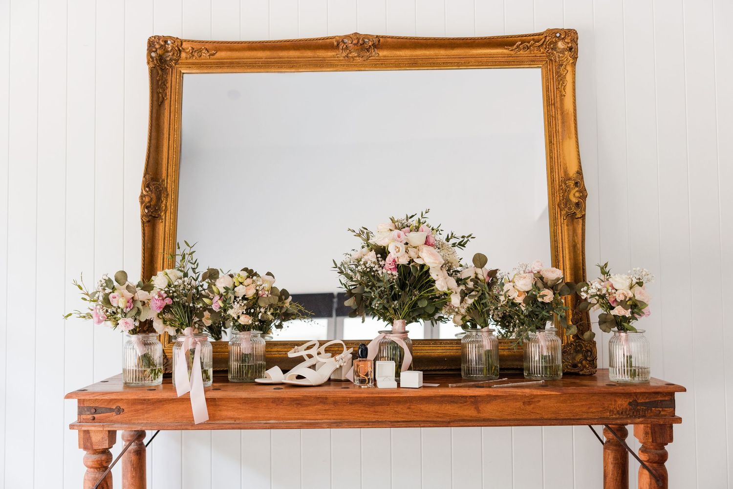 Elegant wooden console table with pink floral arrangements and a large ornate gold mirror against a white wall.