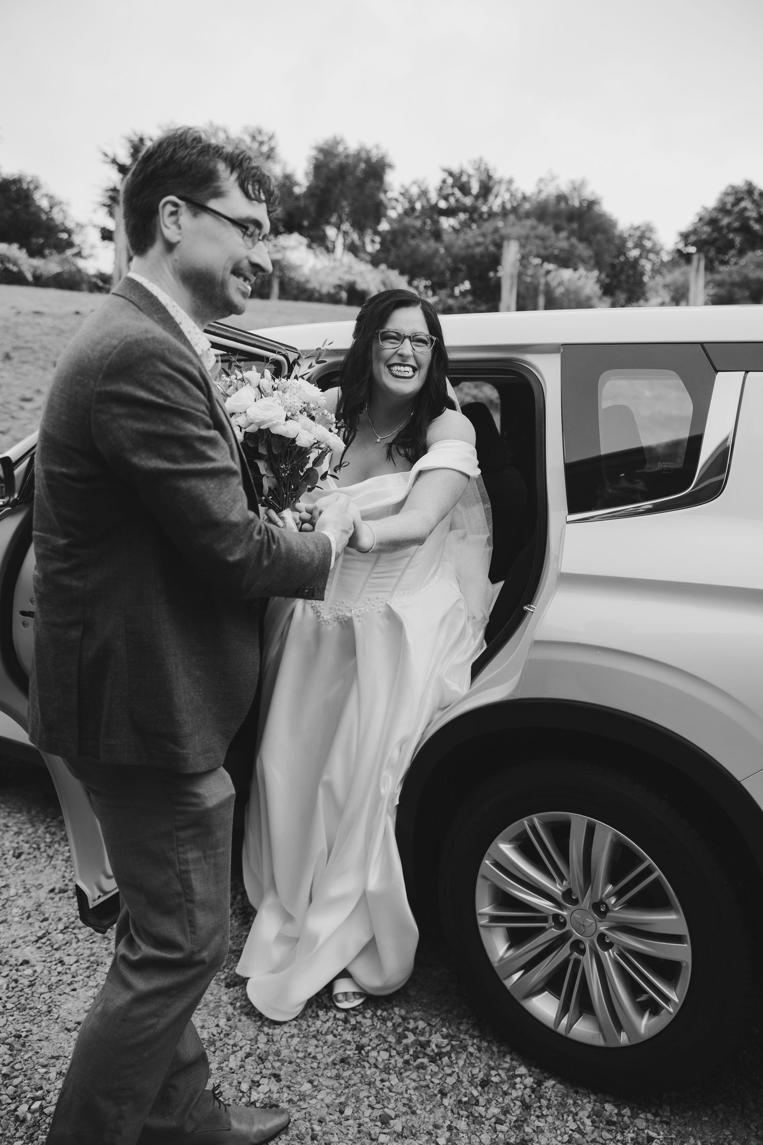 Black and white photo of a laughing bride in white gown being helped out of a limousine by a smiling groom holding flowers.