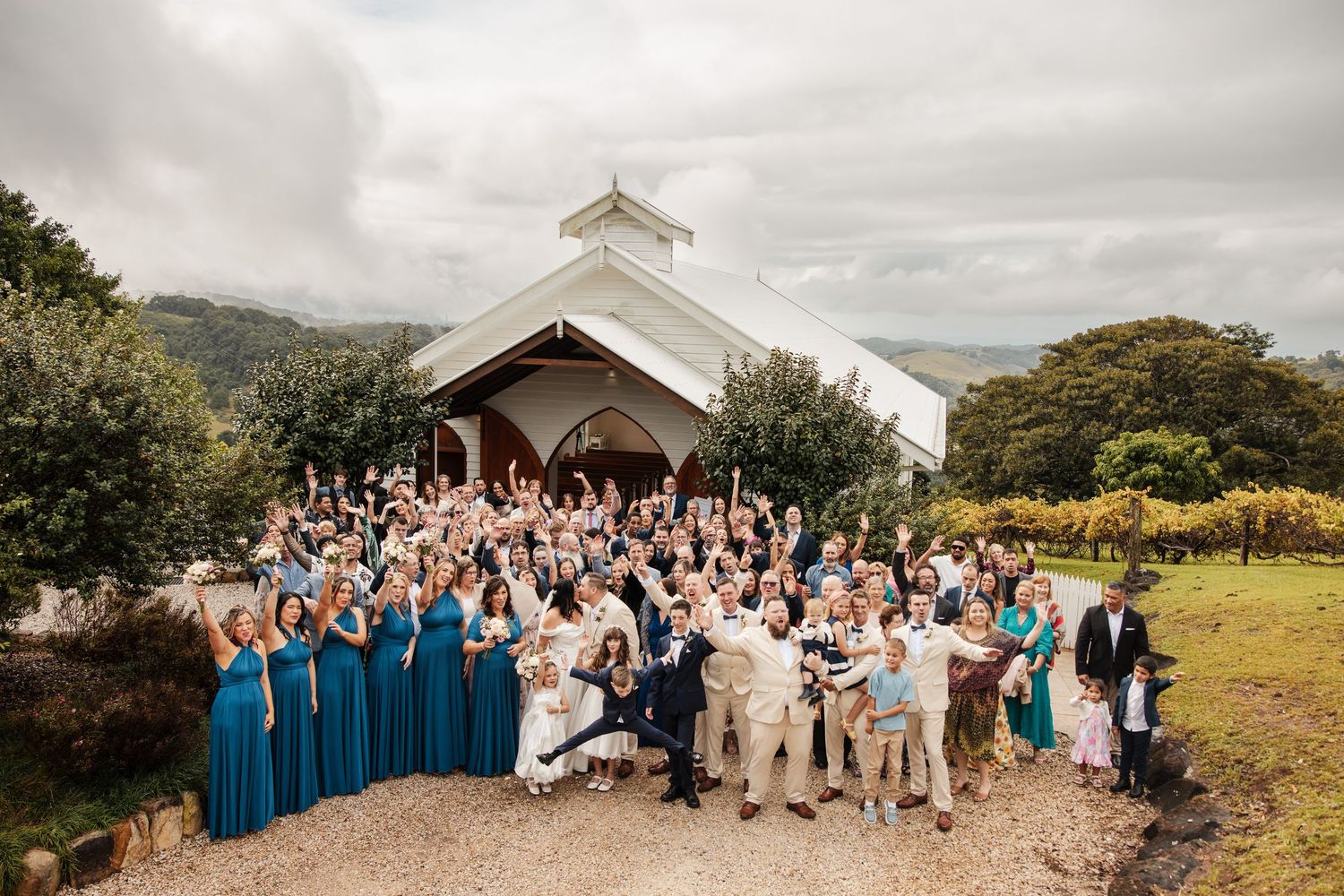 Large wedding party celebrating outside a white chapel with lush green hills and vineyard in the background.