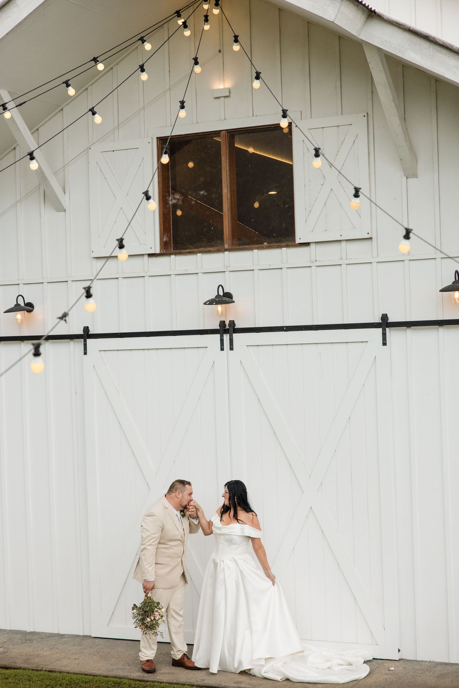 Bride and groom holding hands in front of a white barn with string lights at a rustic wedding venue.