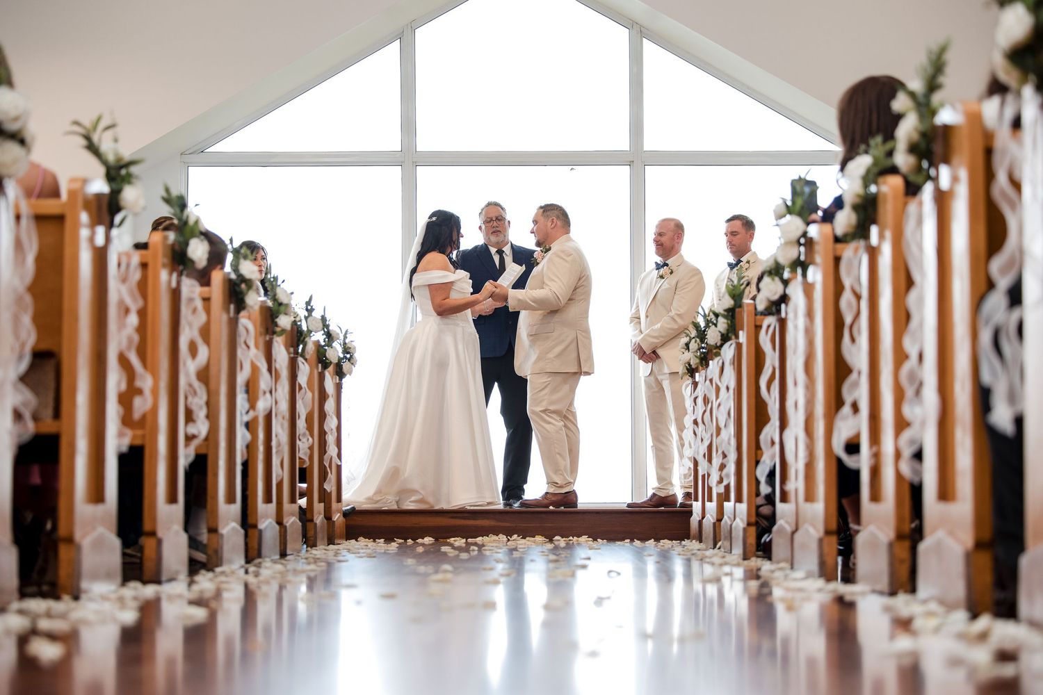 Elegant wedding ceremony in a white chapel with floral-decorated pews, couple exchanging vows before large windows.