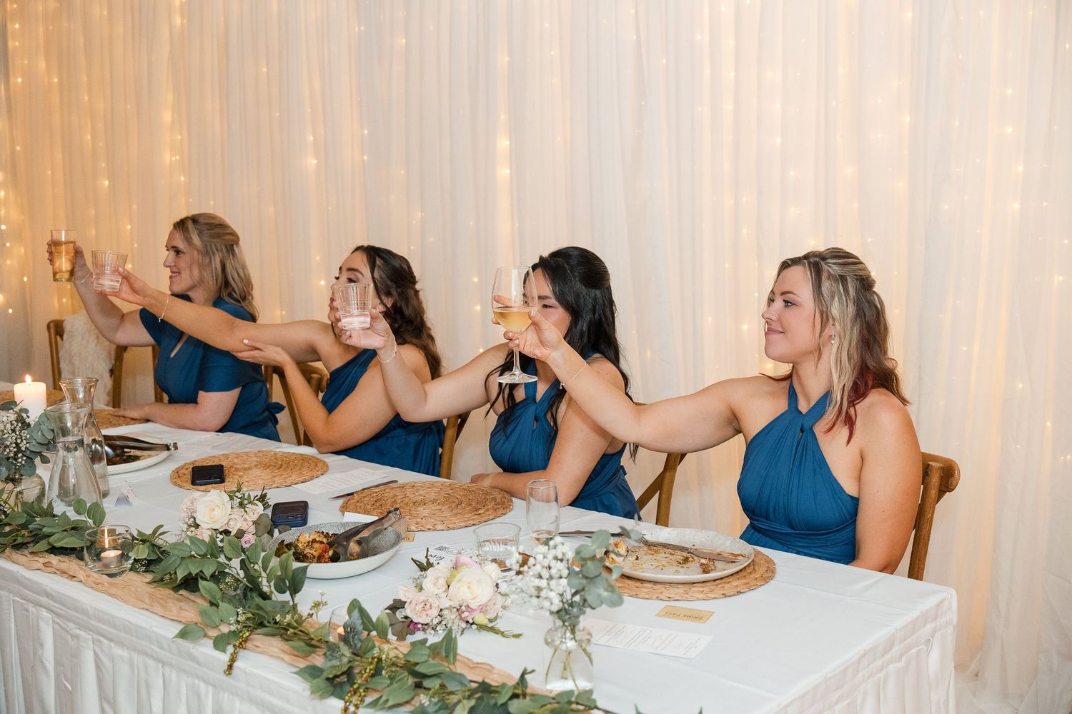 Bridesmaids in blue halter dresses raising champagne glasses at wedding reception table with floral decor.