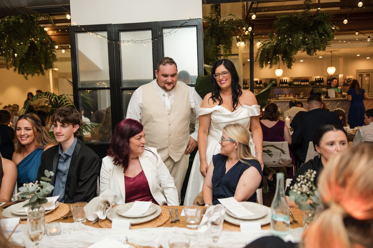 Bride and groom smiling with seated wedding guests at elegantly decorated reception tables in a warmly lit venue.