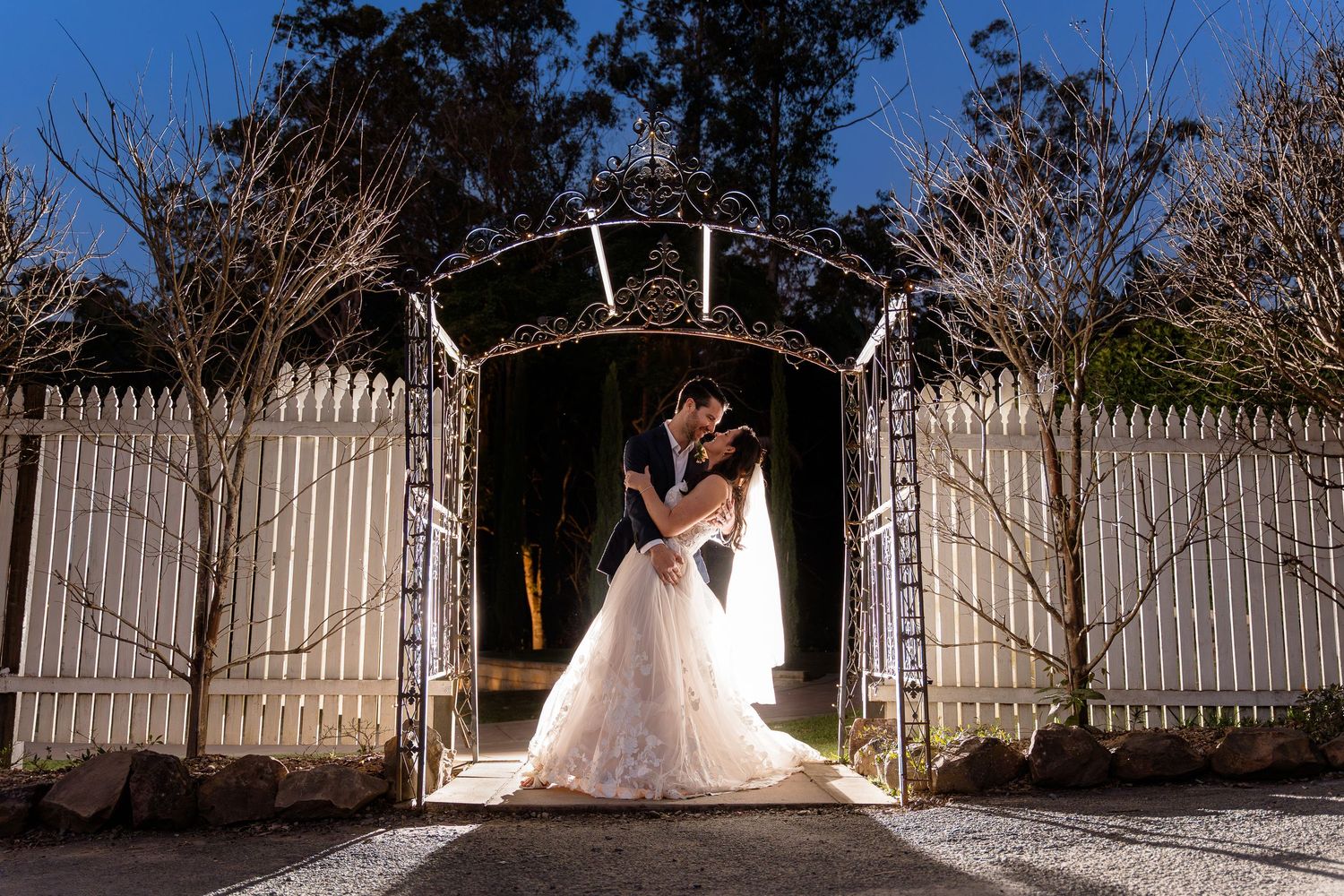 A series of romantic wedding photos taken at night with illuminated white picket fences in the background.