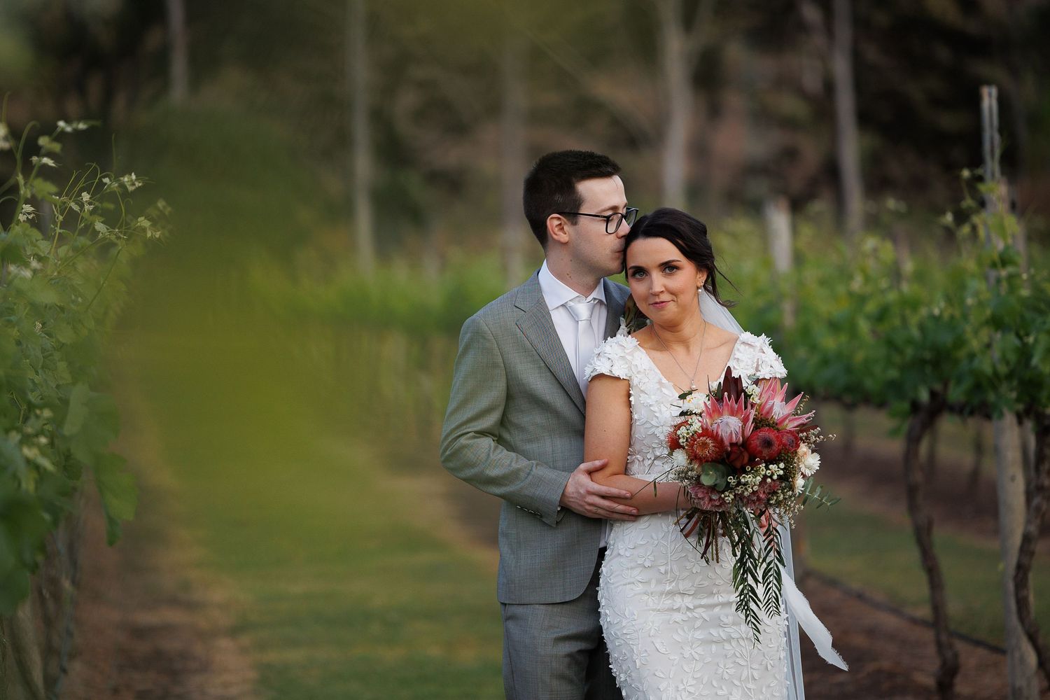 Bride and groom pose romantically in a vineyard, bride holding a protea bouquet, groom in grey suit.