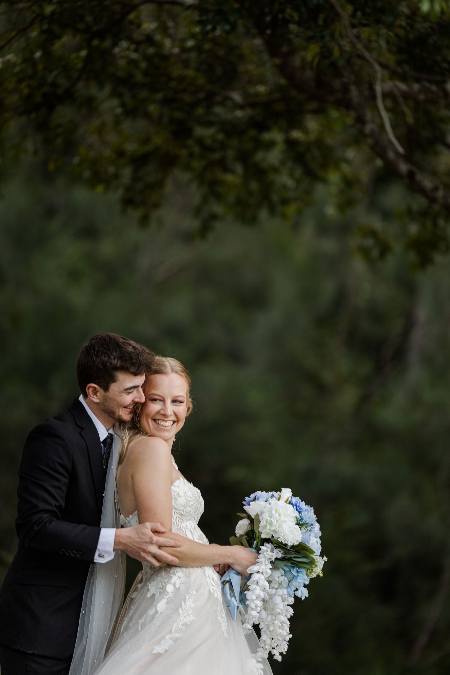 Bride and groom embracing outdoors, she holds blue and white bouquet, wearing lace gown, he in black suit, lush green trees behind.