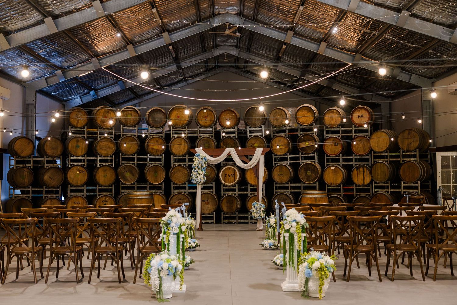 Elegant winery wedding ceremony setup with oak barrels backdrop, wooden cross-back chairs, floral arch, and white aisle flowers.