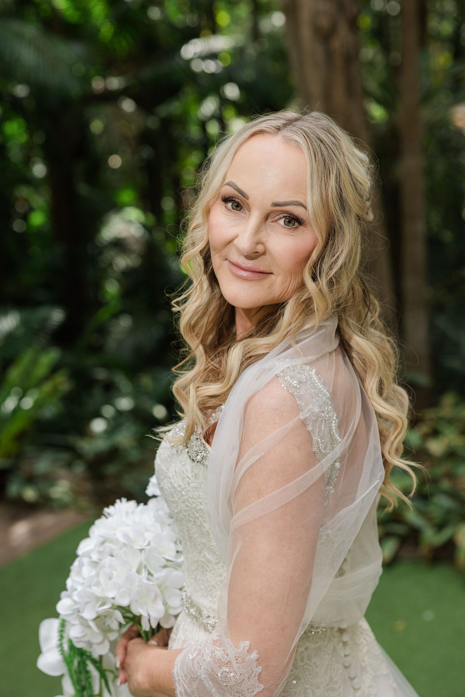 Smiling bride in lace wedding dress holding white bouquet, posing outdoors among lush green tropical foliage.