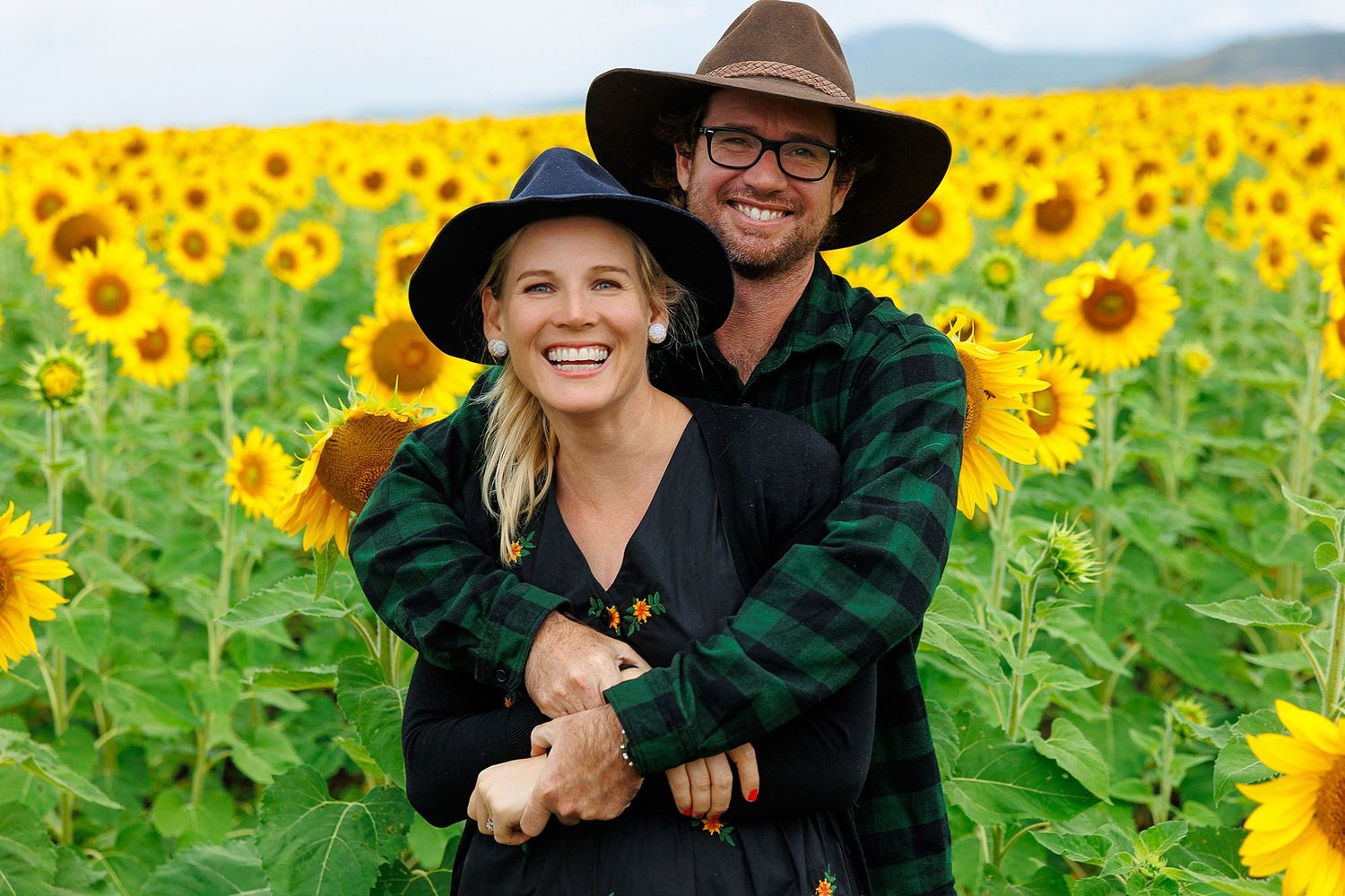 A couple embraces while standing in a bright yellow sunflower field on a sunny day.