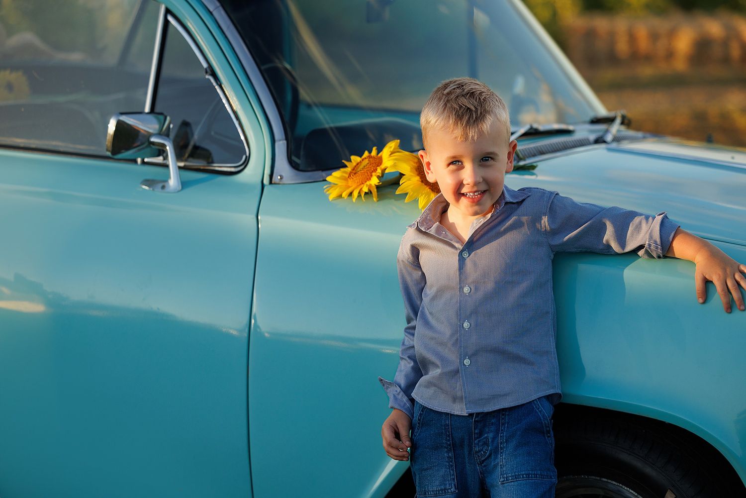 Child leaning against vintage blue car with sunflower at sunset, wearing blue striped shirt.