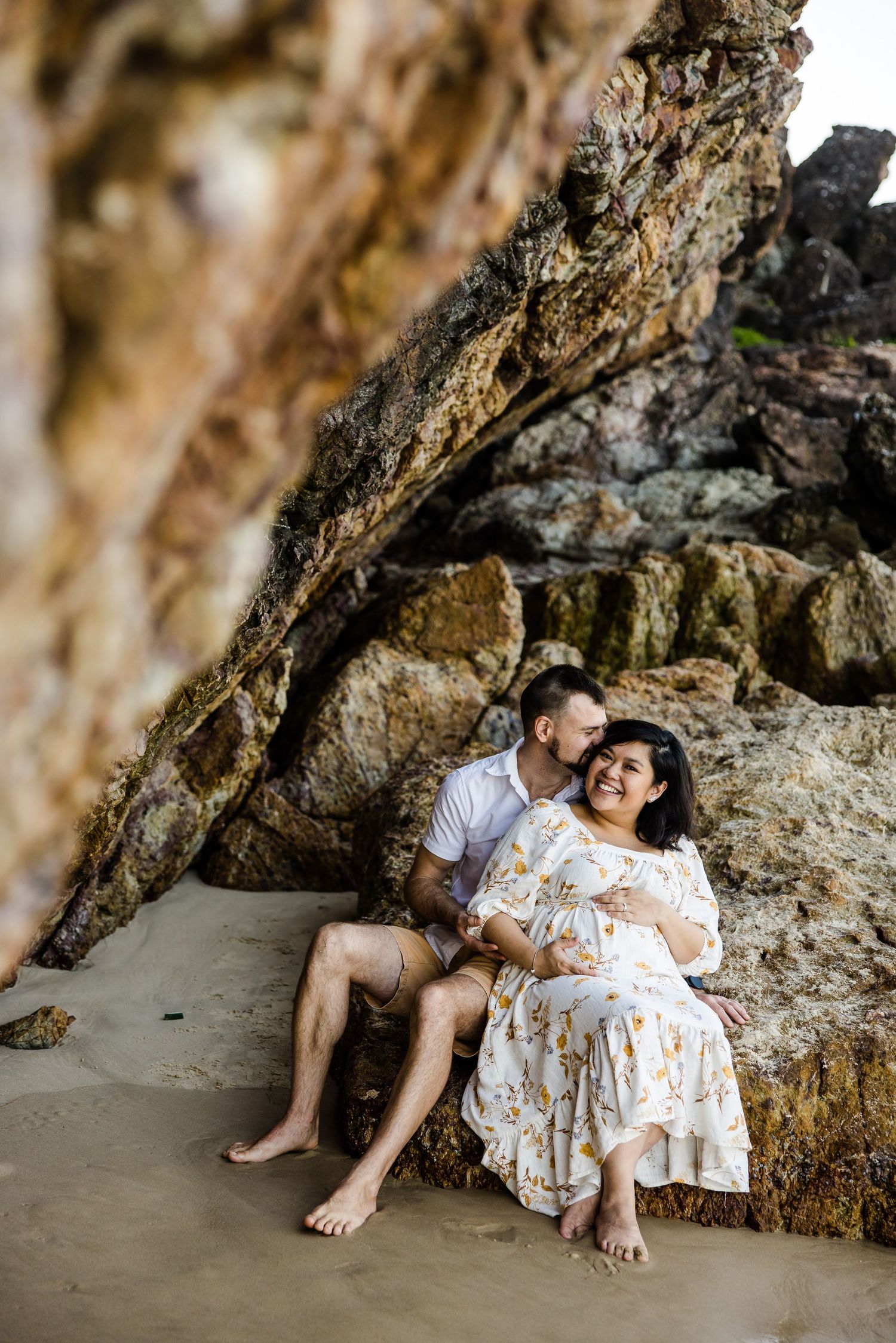 Couple sits together in romantic beach cave setting with dramatic rocky backdrop and diffused natural lighting.