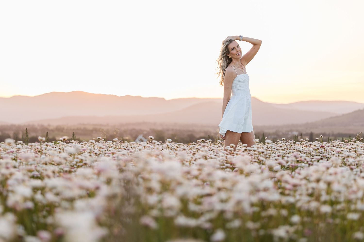 Woman in white dress standing in a field of white daisies at sunset with mountains in the background.