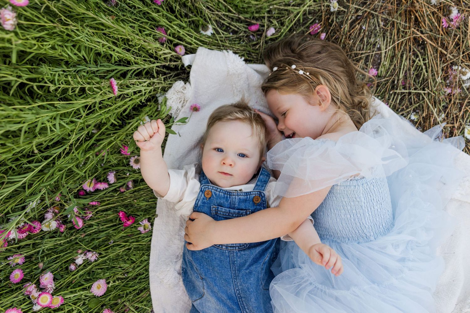 Two young sisters lying in a field of pink wildflowers, older girl in blue dress cuddling baby in denim overalls.