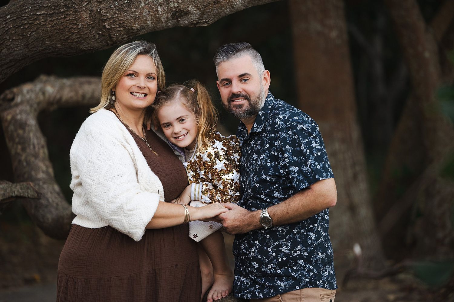 A warm outdoor family portrait shows three people embracing and smiling in front of a rustic backdrop.
