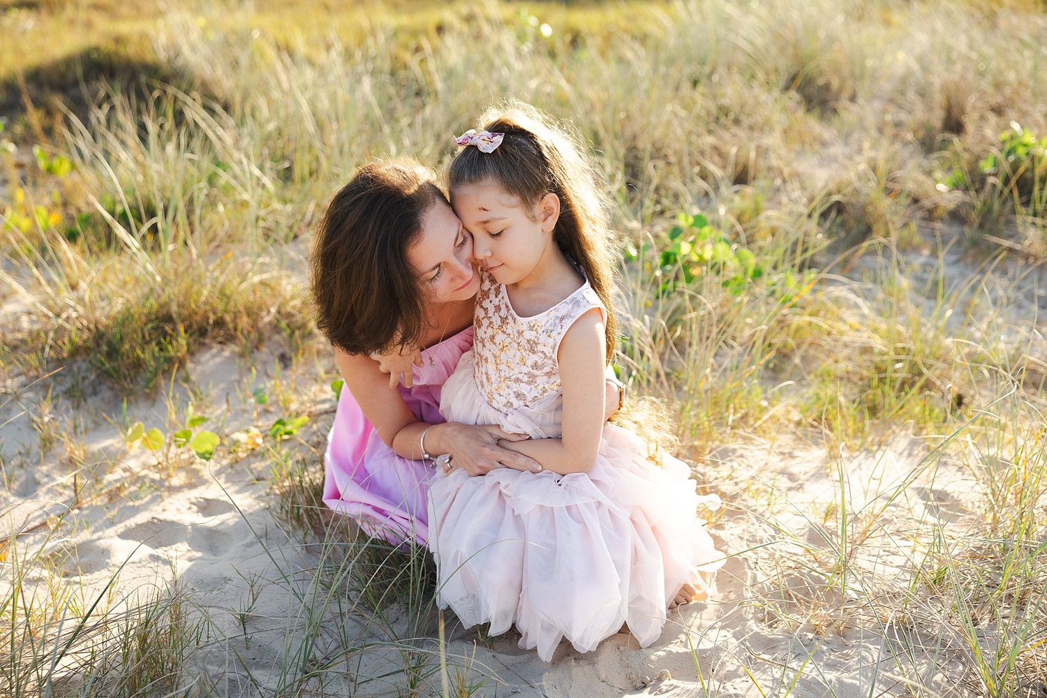 Two people in pink dresses sit close together on sandy ground surrounded by tall grass in warm sunlight.