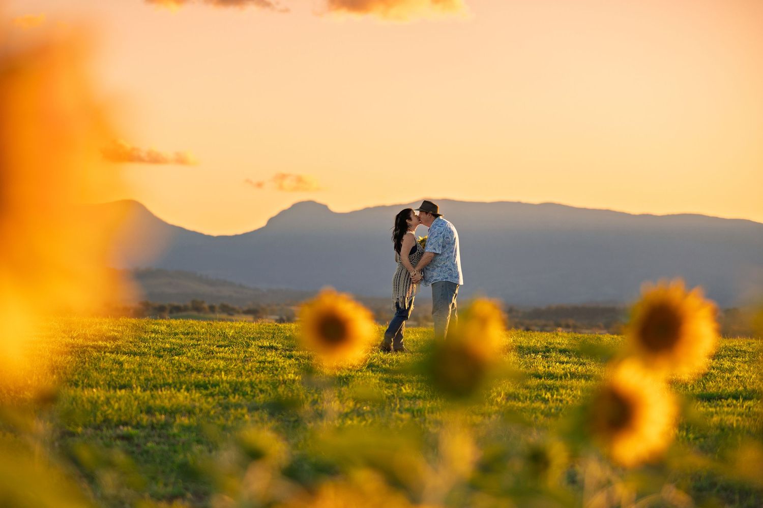 A silhouetted couple embraces in a field at sunset with sunflowers in the foreground and mountains in the distance.