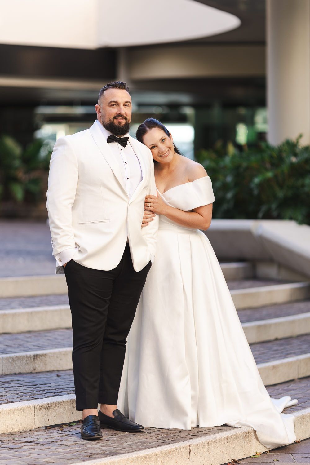 A wedding couple poses on stone steps, with one wearing a white tuxedo jacket and black pants and their partner in a white gown.