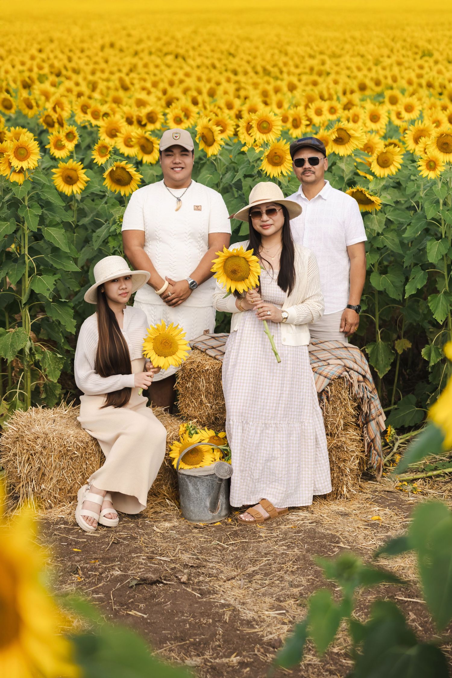 Family sits on hay bales in a vibrant sunflower field wearing coordinated white and beige outfits.