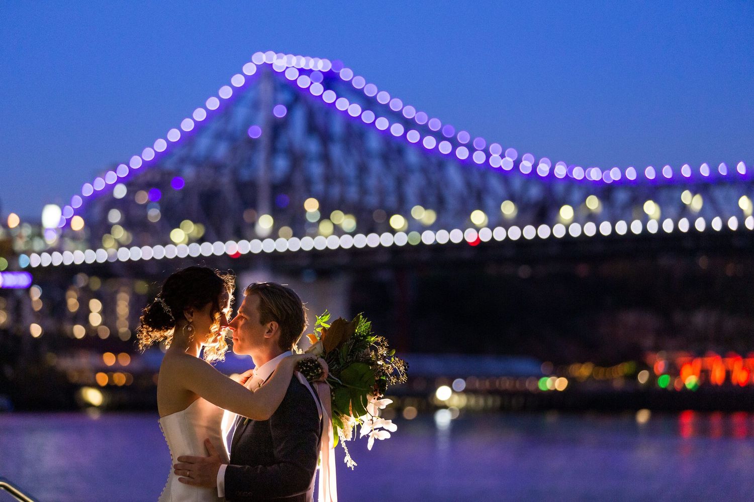 A romantic evening scene shows a couple silhouetted against a bridge illuminated by twinkling lights at dusk.