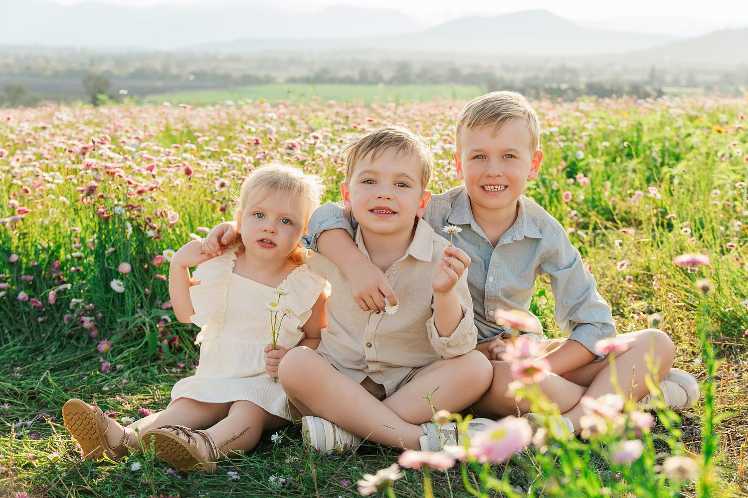 Three smiling children sit close together in a blooming wildflower meadow on a sunny day with mountains in the background.