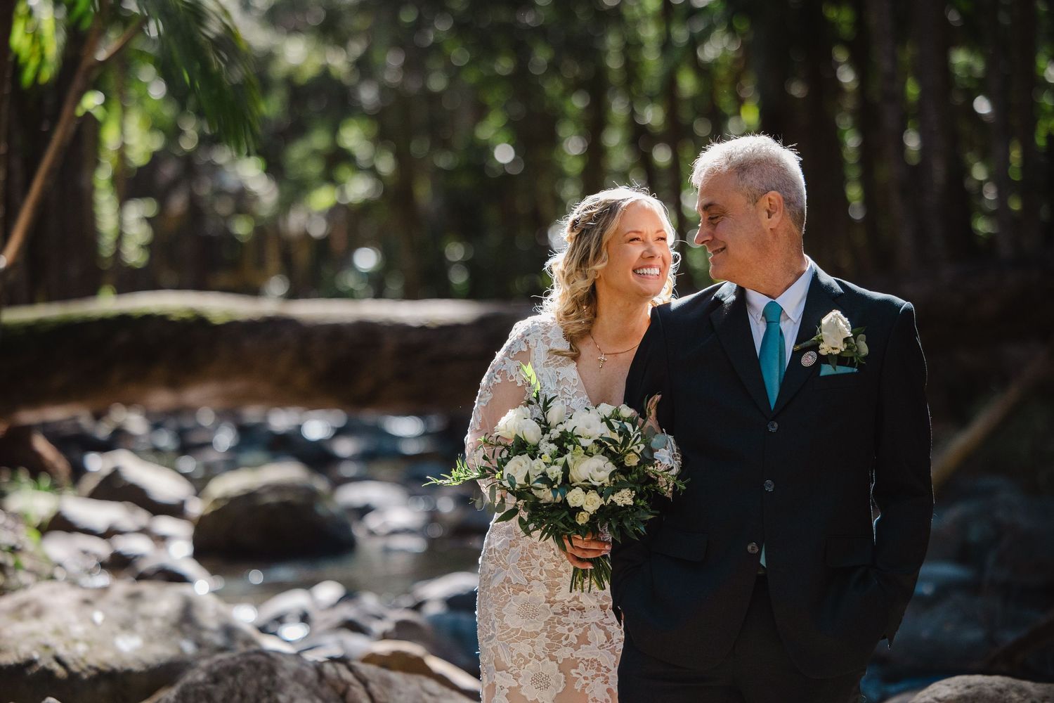 Bride and groom share a joyful moment outdoors near a rocky stream, surrounded by lush forest trees.