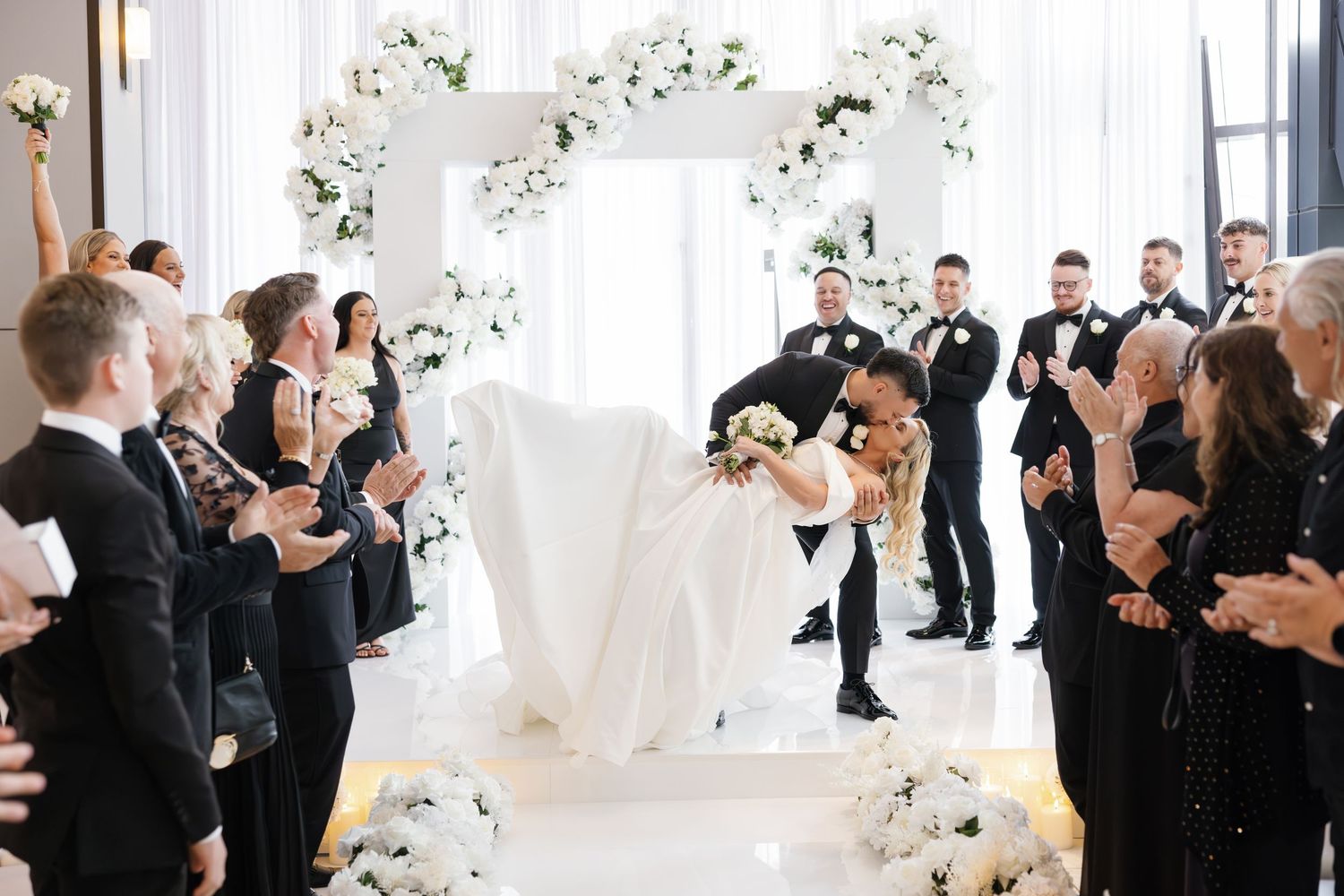 Wedding guests throw white confetti in celebration during an elegant indoor ceremony with bright decor.