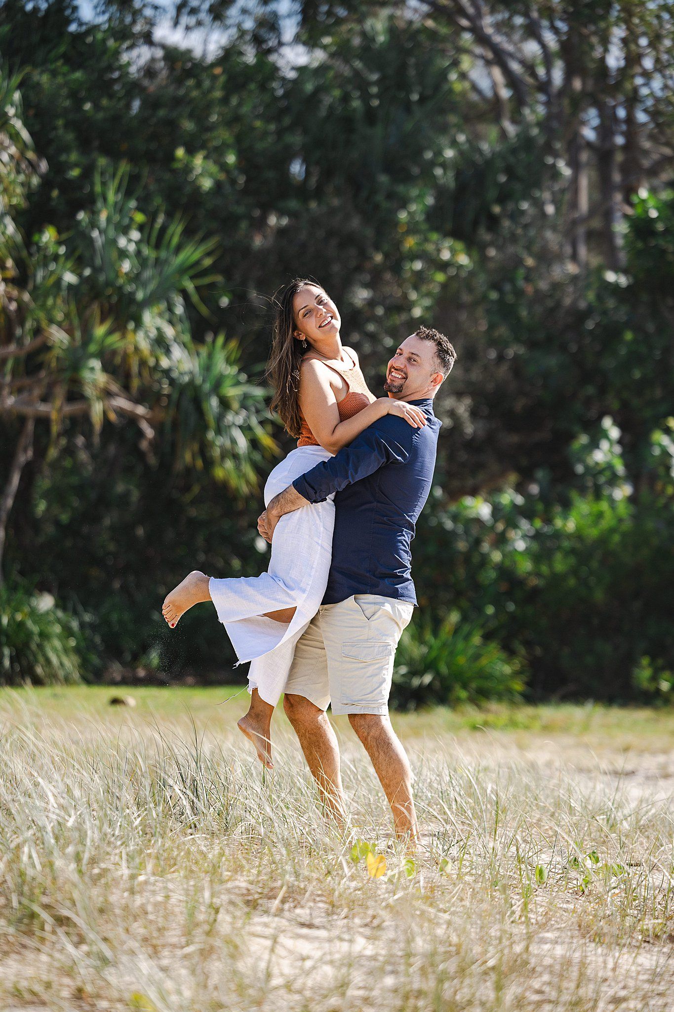 A couple shares a joyful moment in a sun-drenched field as they embrace and laugh together among tall grass and trees.