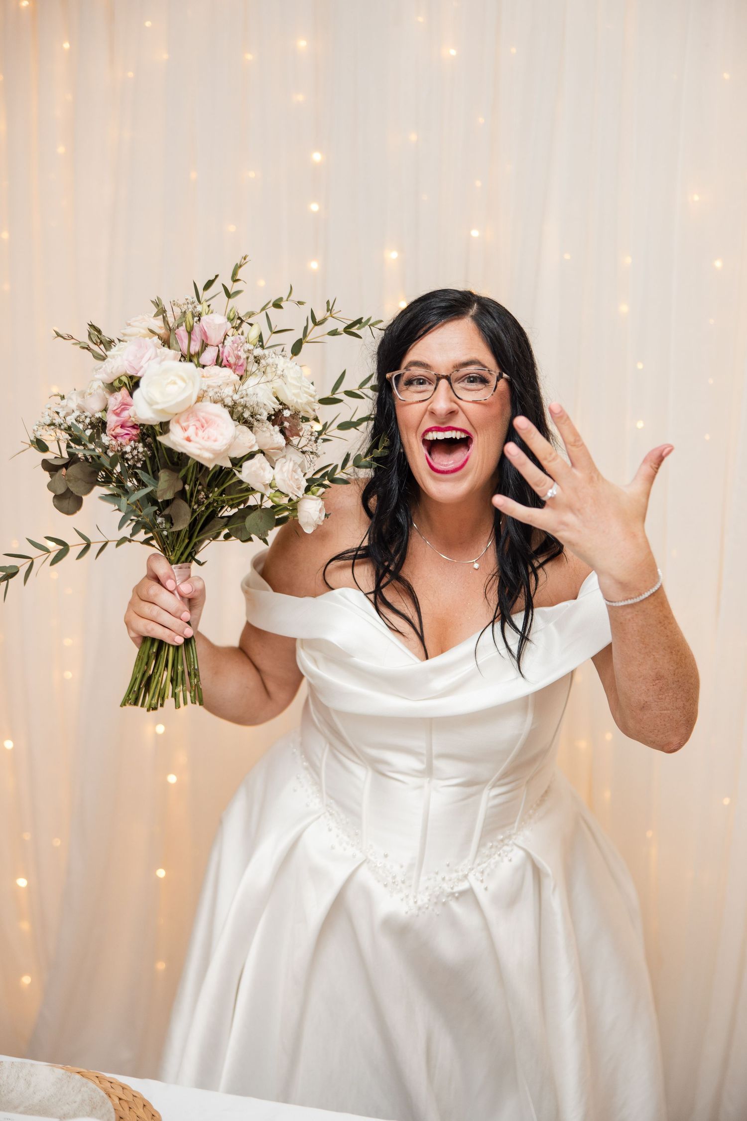 Excited bride in white off-shoulder gown showing engagement ring while holding pink floral bouquet against fairy light backdrop.
