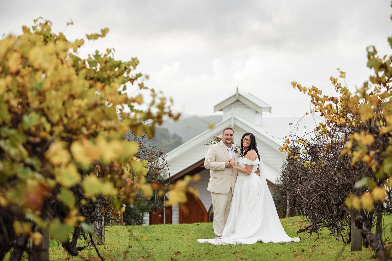 Bride and groom pose in a lush vineyard with a white chapel backdrop on their wedding day.