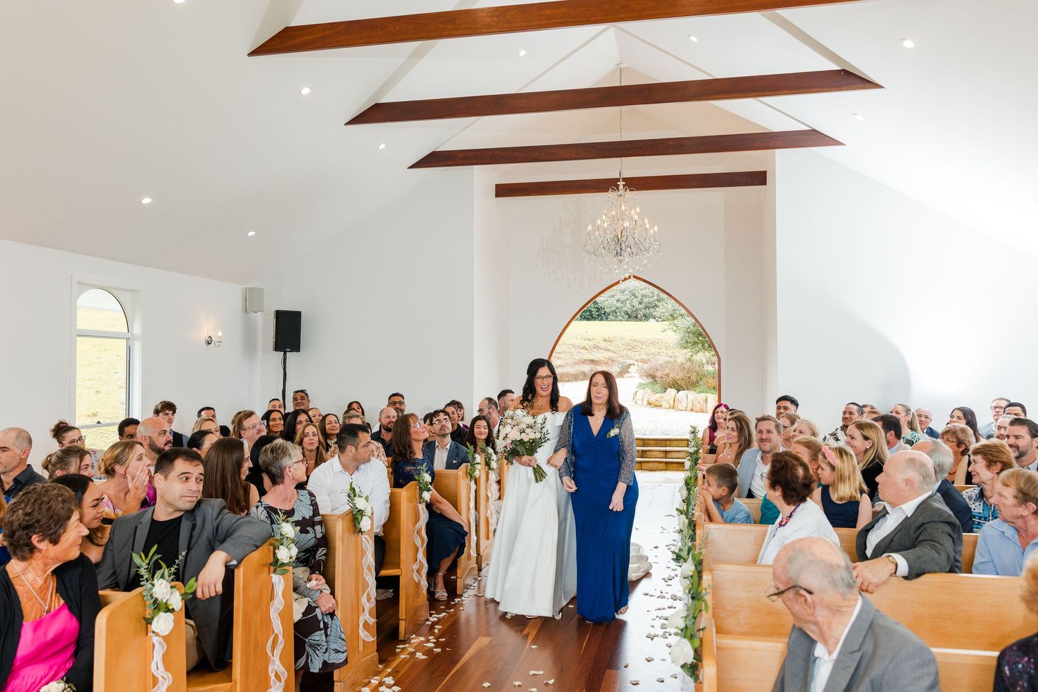 Bride walks down the aisle in a white chapel wedding ceremony with guests seated in wooden pews.