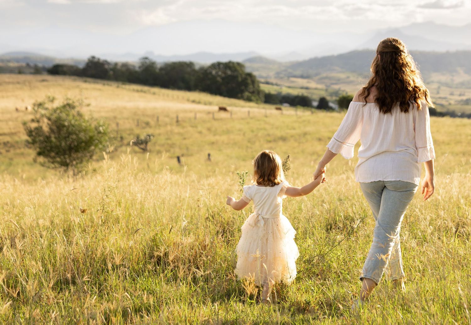 Two people walking hand in hand through a sunlit golden meadow with rolling hills in the background at sunset.