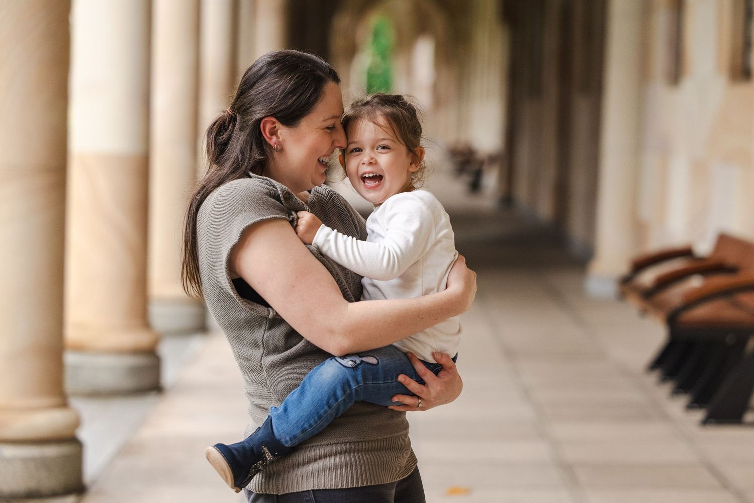 A parent and child share a joyful moment dancing together in a corridor with elegant stone pillars.