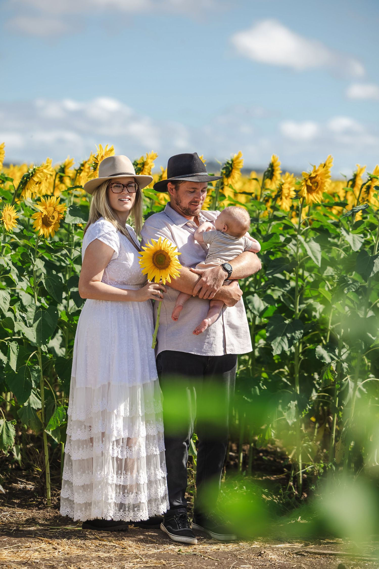 Young couple in hats holding their newborn baby in front of a lush sunflower field.