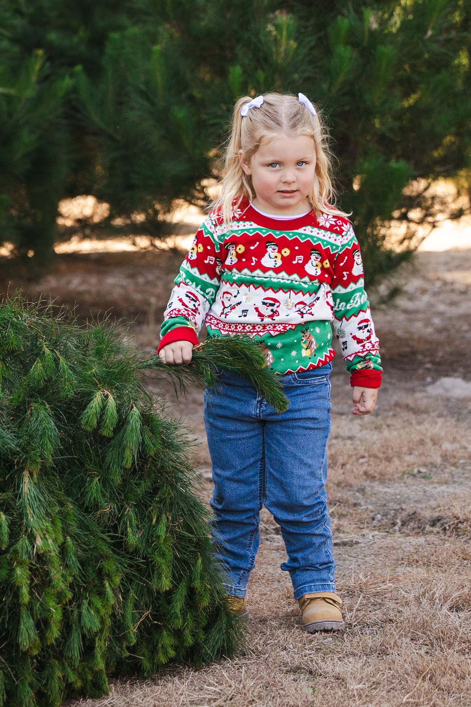 A child wearing a festive holiday sweater and jeans stands near evergreen branches on brown ground.