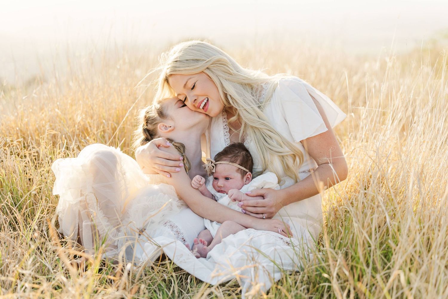 Blonde mother sitting in golden field, tenderly kissing older child while cradling newborn baby in soft natural light.