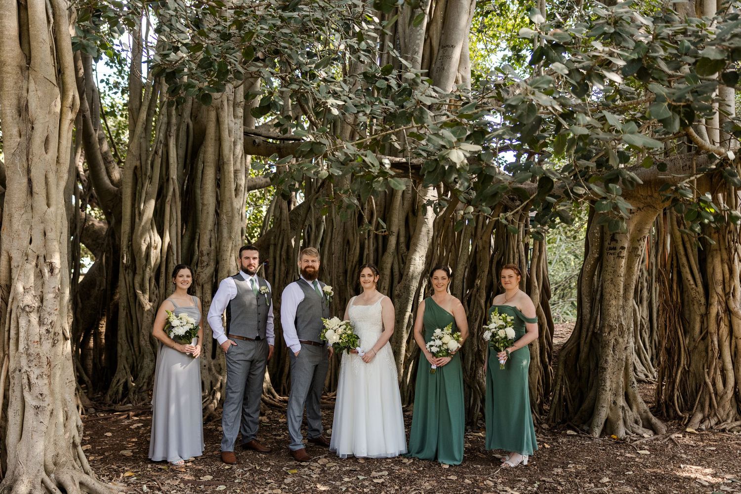 Wedding party posing under a large banyan tree, bridesmaids in gray and green gowns with floral bouquets.