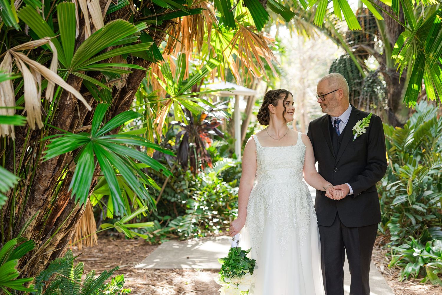 Bride in white gown walks with older gentleman through lush tropical garden path on wedding day.