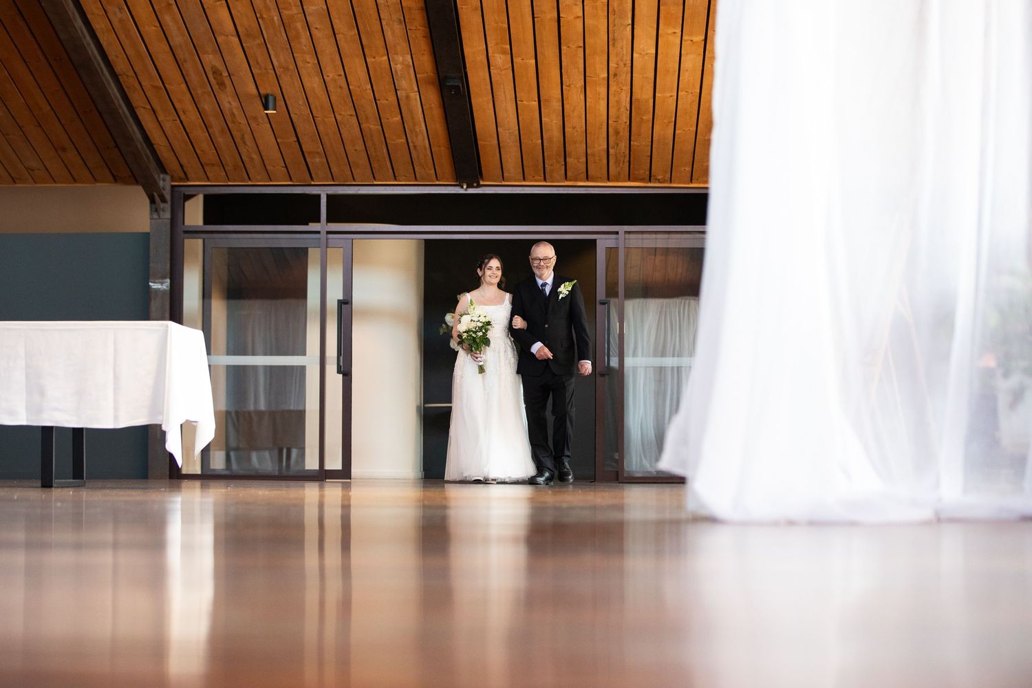 Bride and groom standing together at wedding venue entrance with wooden ceiling and sheer white curtains.