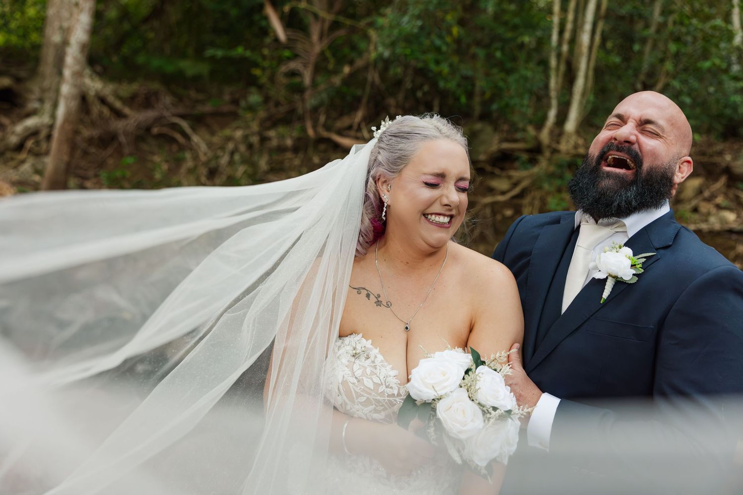 Joyful bride and groom laughing outdoors, brides veil flowing in wind, groom in navy suit, bride holding white bouquet.