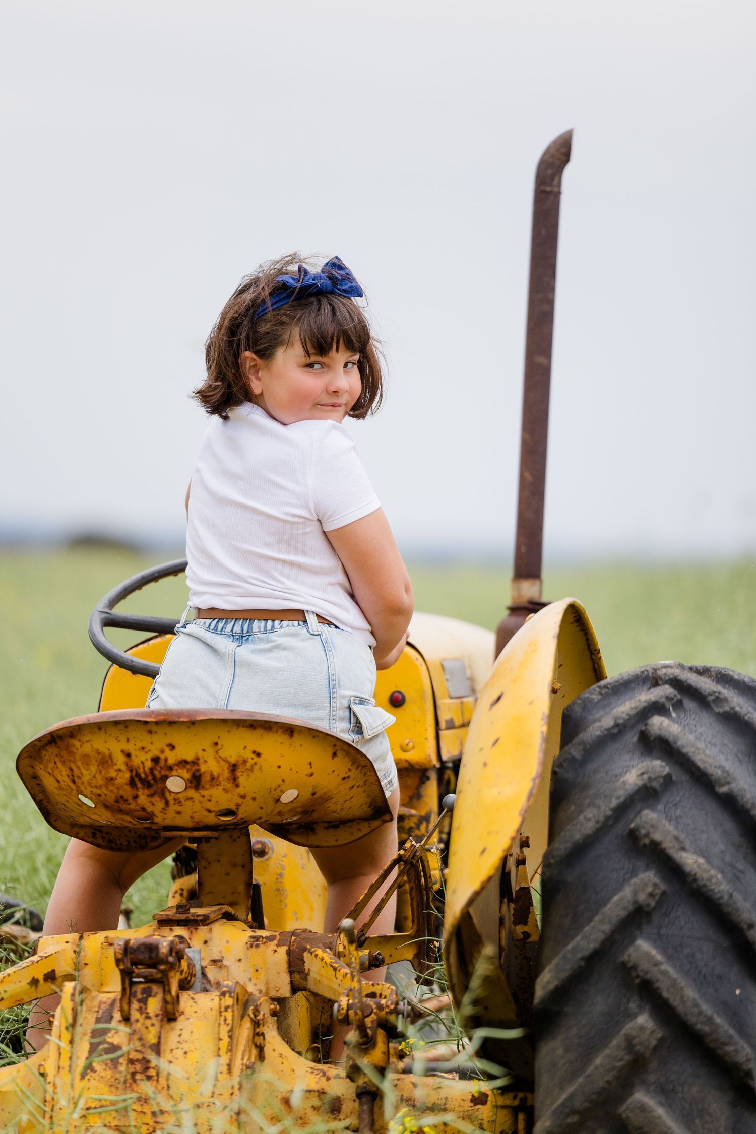 Young girl in white top and denim shorts sitting on a vintage yellow tractor in a green field.