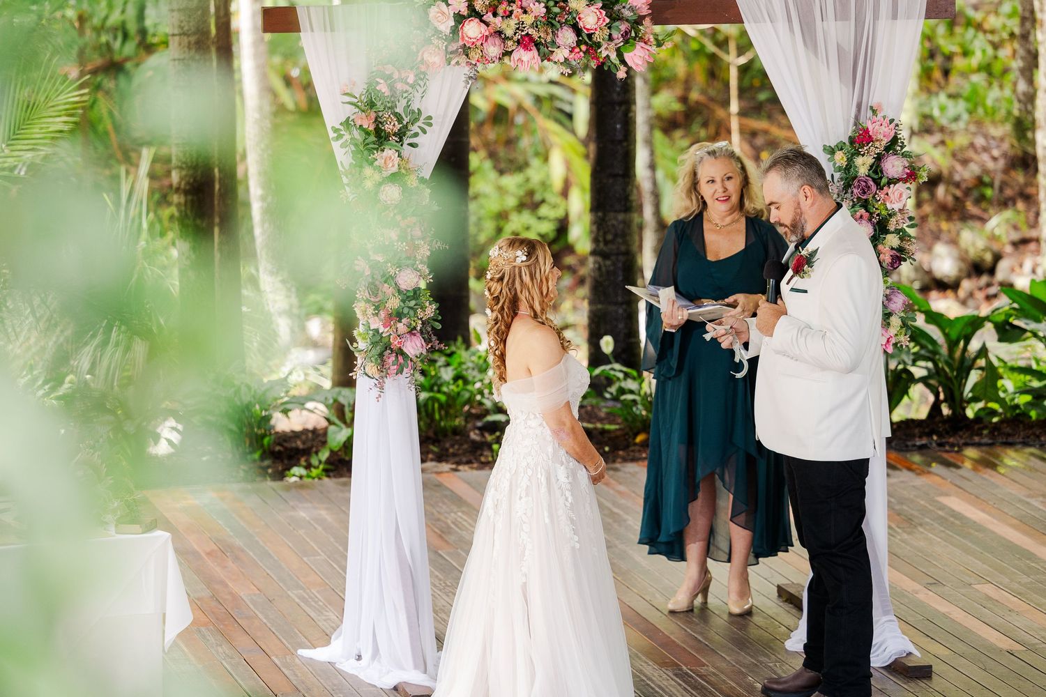 Bride and groom exchange vows at an outdoor wedding ceremony with pink floral arch and draped white curtains.