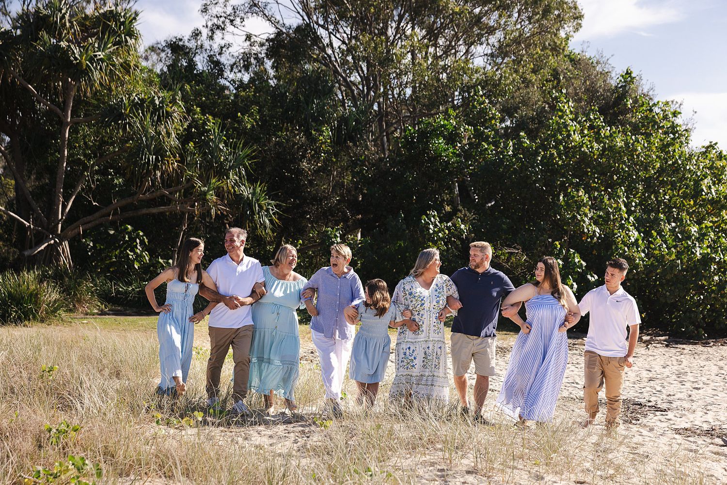 A group dressed in soft blue and white coordinated outfits walk together on a beachside path surrounded by greenery.